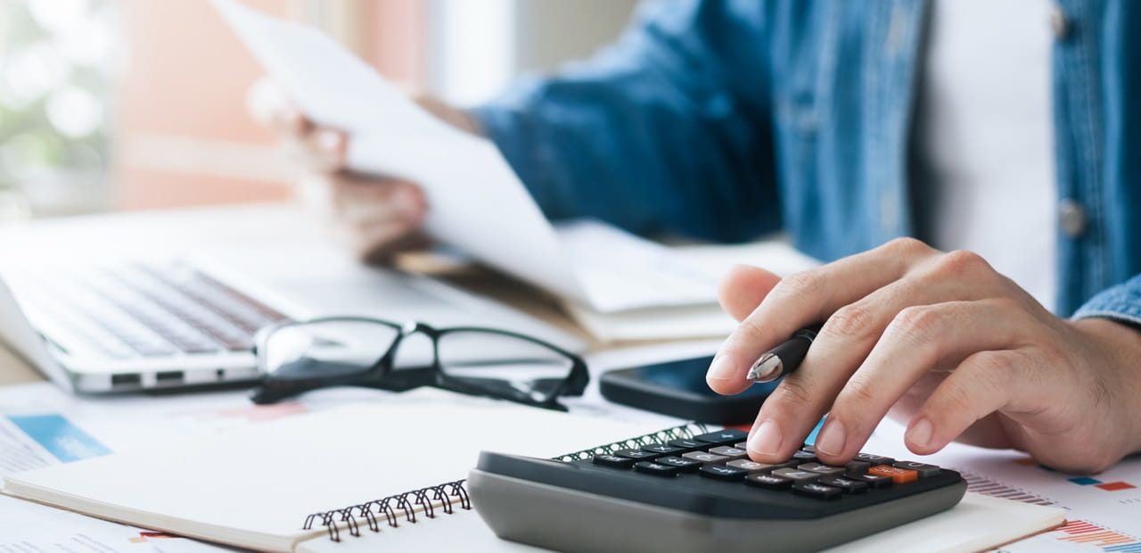 Close-up of a person using a calculator at a desk with documents, a laptop, eyeglasses and a smartphone.