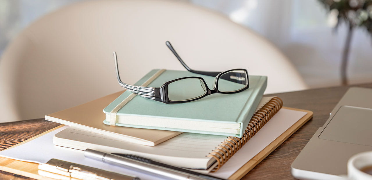 Stack of notebooks and papers on a desk with eyeglasses, a pen, and part of a laptop.