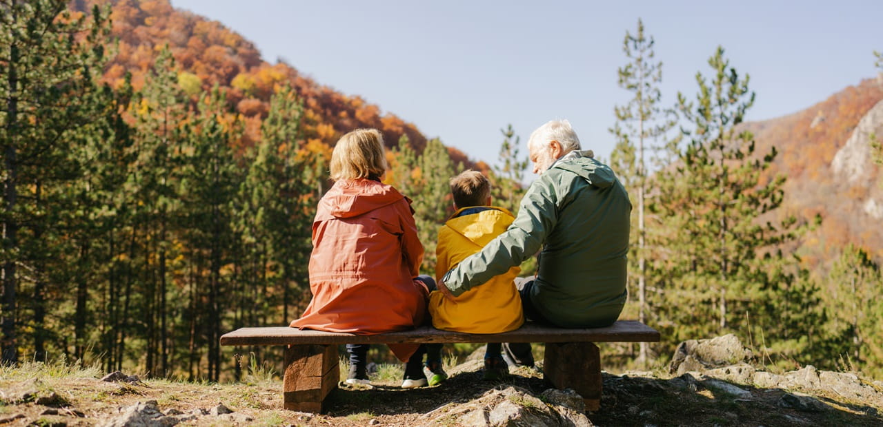 Grandparents sitting on a bench with their grandchild, overlooking a forested mountain with autumn colors under a clear sky.