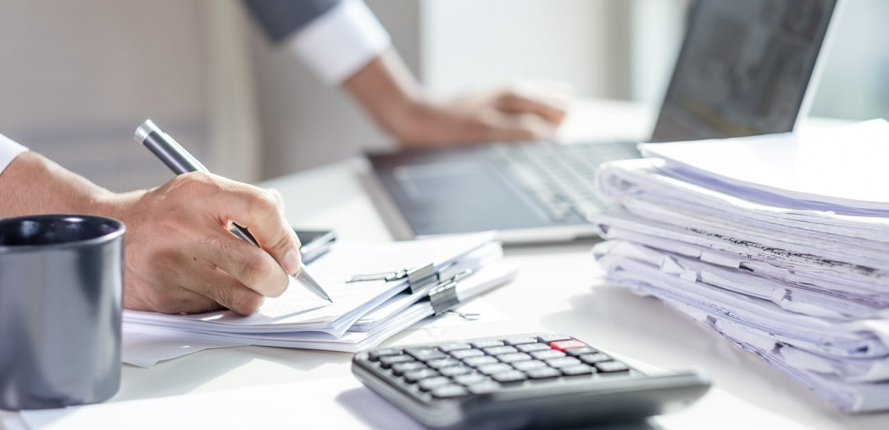 Person writing on documents at a desk with a calculator, coffee mug, laptop, and a stack of papers.