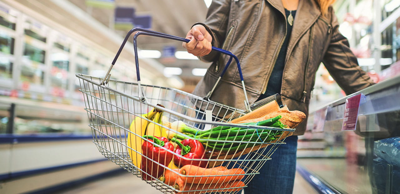 Person holding a shopping basket filled with organic produce in a grocery store aisle.