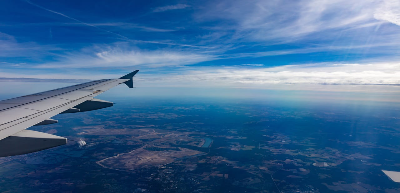 View from airplane window showing wing over a landscape with scattered fields and rivers under a blue sky with wispy clouds.